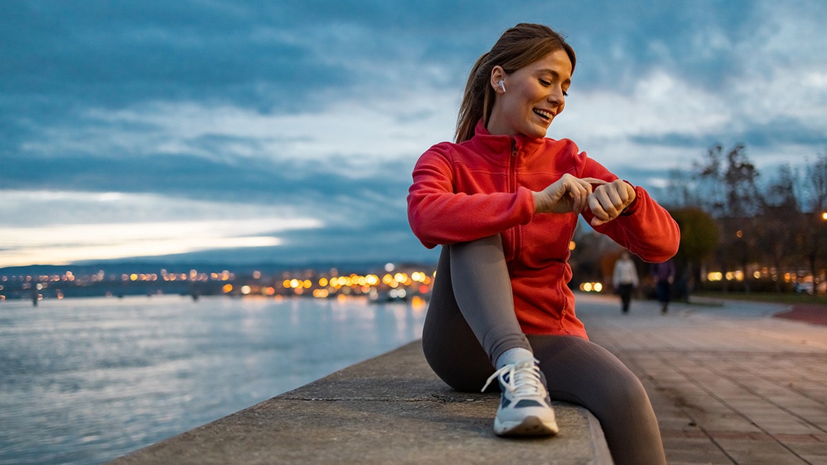 The woman with running equipment checks the heart rate with a smart watch. It wears a red long sleeve zipper and sits on the edge of the water.
