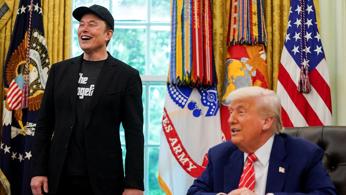 Musk in black at the oval office, next to Trump sitting